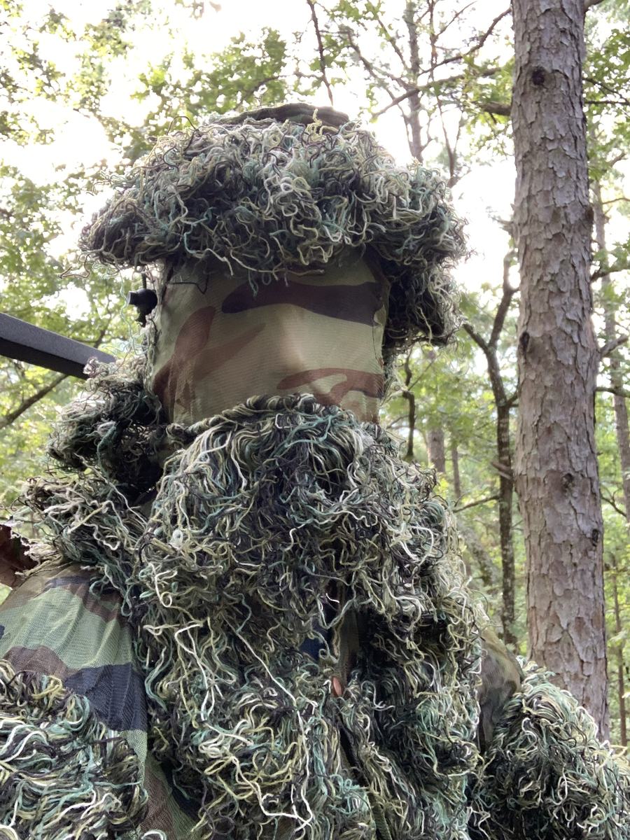A hunter in a ghillie suit and hooded face mask in the pines