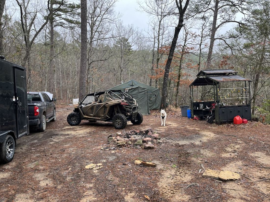 A side-by-side parked at a wooded camp next to a truck and trailer